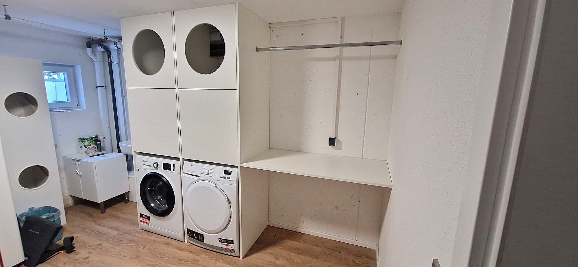 Waschmaschinenschrank nach Maß: 2 Wäschepuffer mit Einwurf-Bullaugen über Waschmaschine und Trockner. Daneben Arbeitsfläche und Trockenstange.